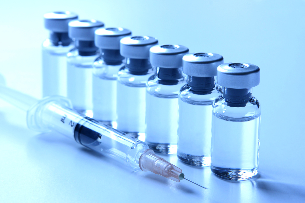 Studio shot of a series of seven vials standing in a row filled with liquid medication. Empty syringe laying next to the vials is ready for injection and  could represent concepts like vaccination, flu shot, medication. Image is blue tinted.